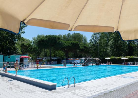 Piscina all'aperto con bagnanti e ombrelloni in una giornata soleggiata.